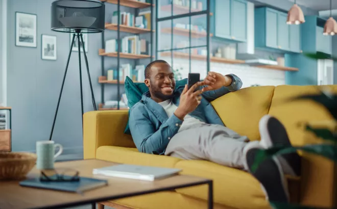 Lächelnder Mann schaut auf sein Smartphone und liegt auf dem Sofa